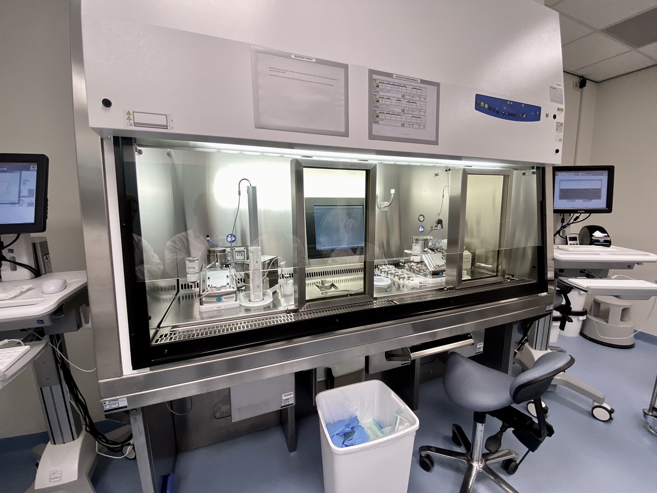 A brightly lit, sterile cleanroom featuring a large, silver and glass radiopharmacy isolator as the central visual element. Workstations with computer screens flank the isolator, creating a sense of a controlled and high-tech environment.