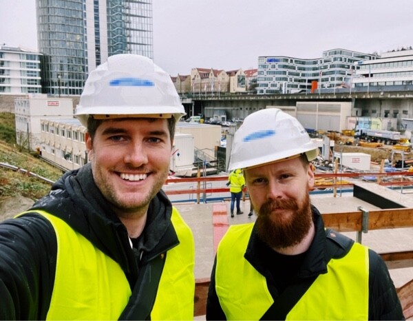A selfie of two visitors, Matthias Nagel and Geffrey van der Bos, wearing white hard hats and yellow safety vests, smiling in front of a construction site. The background shows ongoing construction with buildings and equipment, suggesting they are observing the site.